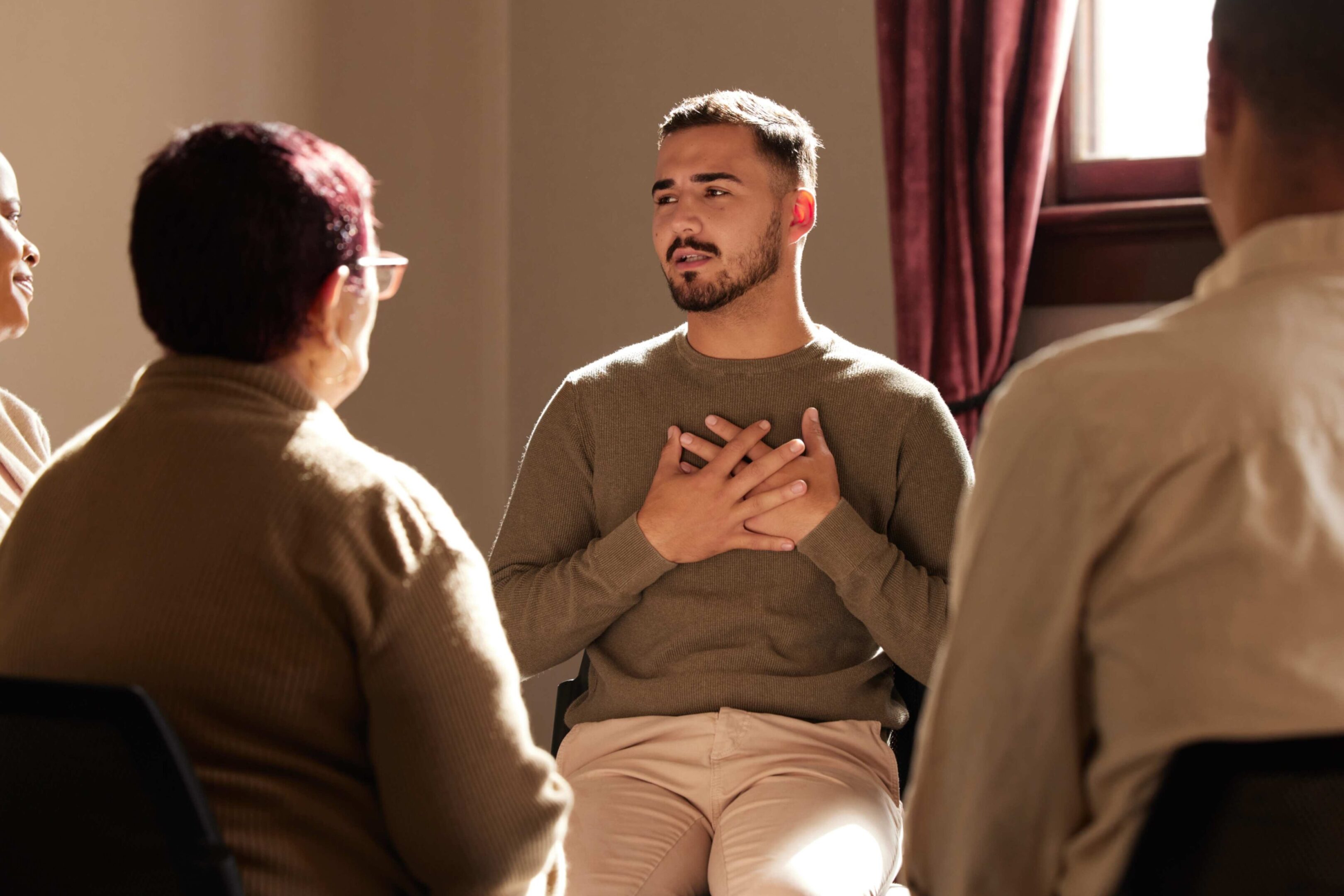 Four people sit in a circle having a discussion; one man in the center holds his hands to his chest while talking.