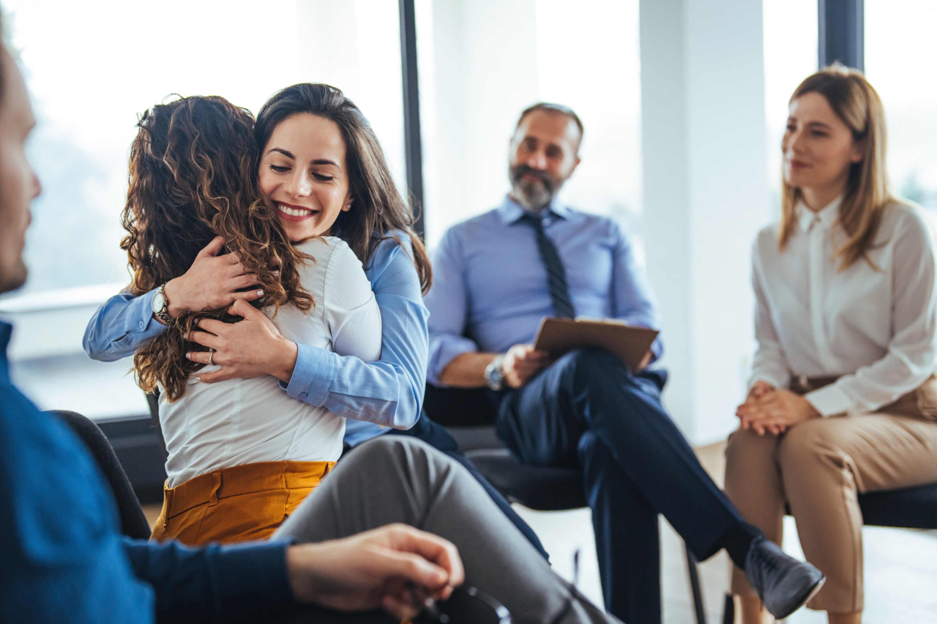 Two women hug while seated in a group therapy session, with two other people watching and one man taking notes.