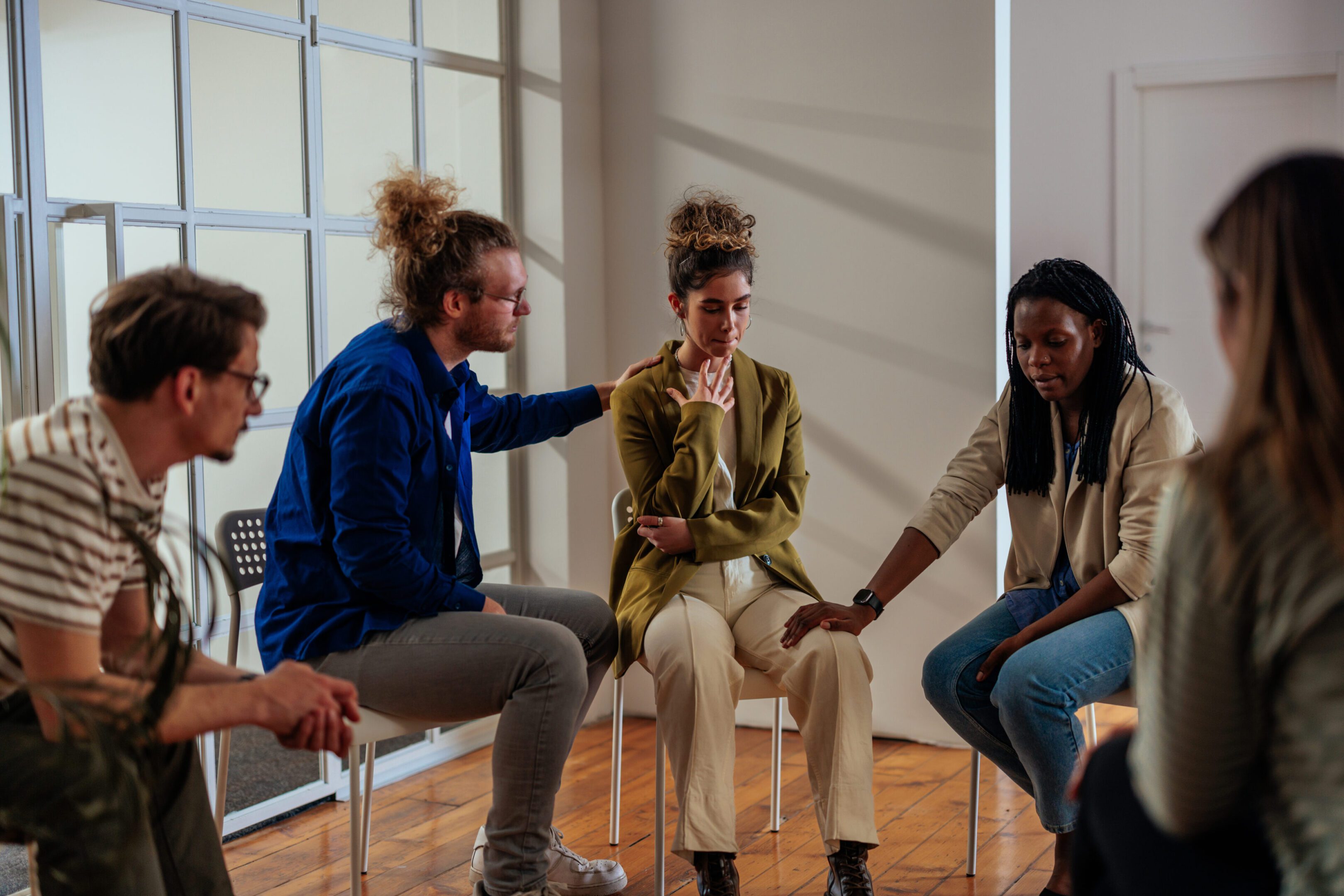 A multiracial group of adults are attending a group therapy session. The attendees are seated in a circle. Young woman shares struggles with the group. Other people around her expressing comfort and support.
