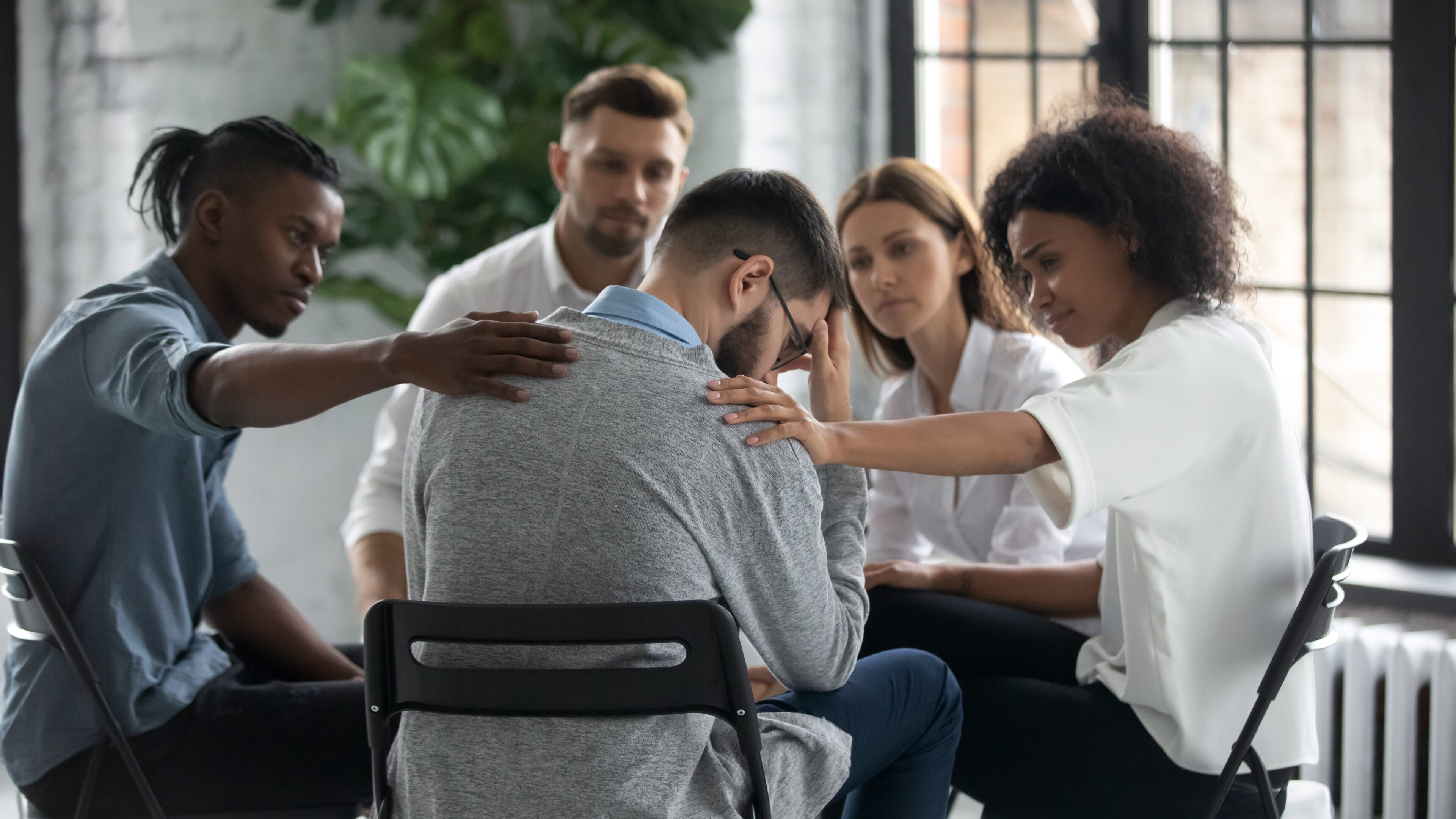 Diverse people sit in circle participate in group psychological session, touch support upset friend, multiracial colleagues comfort unhappy stressed male suffering from mental addiction problems